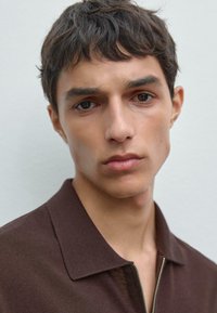 Camicia marrone con zip e colletto, texture a coste. Modello con capelli corti e scompigliati, tono di pelle neutro e mascella ben definita. Lo sfondo è semplice.
