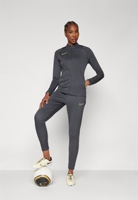 Gray athletic tracksuit with long sleeves, zip front, Nike logo in neon green, slim fit pants, paired with soccer shoes and a ball.