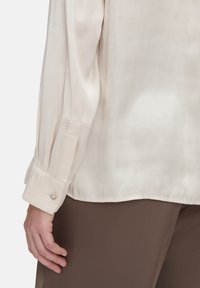 Cremfarbene Seidenbluse mit langen Ärmeln, Knopfmanschetten und einer glatten Textur. Über braunem Stoff getragen, der einen subtilen Glanz zeigt.