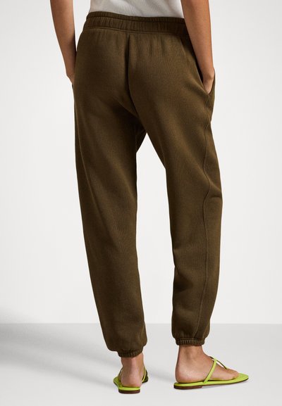 Brune avsmalnende joggebukser med elastisk midjebånd, sidelommer og ribbestrikkede mansjetter, kombinert med limegrønne sandaler. Glatt stofftekstur.