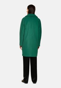Grüner Wollmischungs-Mantel mit Reverskragen, langen Ärmeln und geradem Schnitt, getragen über schwarzen Hosen, von hinten gezeigt.