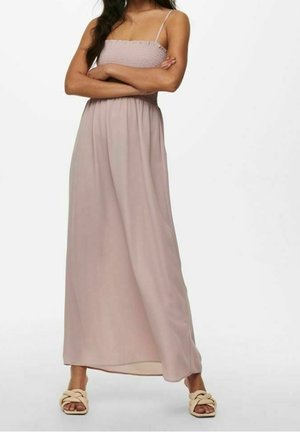 Femme aux longs cheveux foncés portant une robe longue bustier mauve clair et des sandales tissées crème, debout les bras croisés.