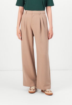 Pantalons beige à jambes larges avec plis sur le devant, portés avec des baskets à imprimé animal et un haut à manches courtes vert foncé, se tenant devant un fond uni.