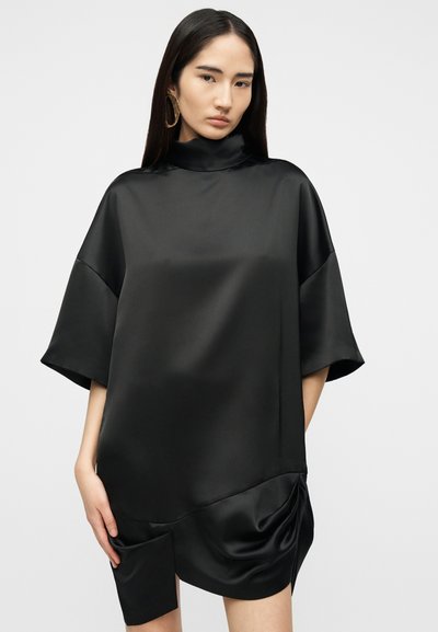 Γυναίκα με μακριά σκουρόχρωμα μαλλιά, φορώντας ένα φαρδύ μαύρο σατέν φόρεμα με φαρδιές μανίκες και ασύμμετρο στρογγυλεμένο τελείωμα, που στέκεται μπροστά σε λευκό φόντο.