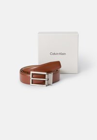 Brązowy skórzany pasek z prostokątną srebrną klamrą. Zawiera logo na klamrze i jest prezentowany obok białego pudełka z napisem "Calvin Klein."