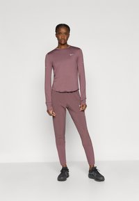 Πλεκτό πουκάμισο με μακριά μανίκια και εφαρμοστά παντελόνια σε μωβ απόχρωση, κατασκευασμένα από απαλό ύφασμα. Διαθέτουν το λογότυπο της Nike σε λευκό στο στήθος και στο αριστερό μανίκι.