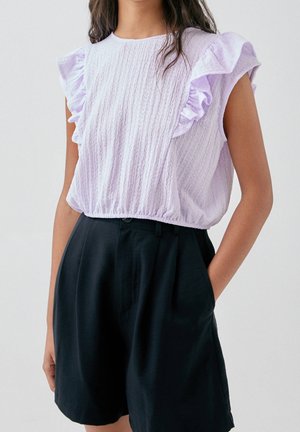 Femme portant un haut sans manches violet clair avec des volants aux épaules et un tissu texturé, associé à un short noir taille haute avec des poches.