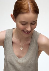 Złota zawieszka w kształcie serca na delikatnym łańcuszku, noszona na beżowej bluzce bez rękawów, podkreślająca jej skomplikowany design i blask.