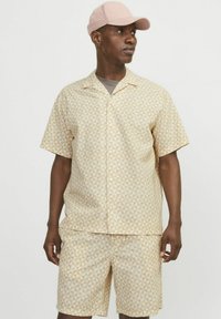 Kurzarm-Hemd mit Knöpfen und passender Shorts in Hellbeige mit geometrischem Muster. Strukturierten Stoff mit lockerer Passform. Pinke Kappe.