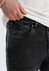 Svarta denimjeans med slim fit, med subtila blekningar, framfickor och metallknappsstängningar. Texturerat tyg synligt nära midjan.
