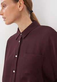 Burgundy blouse with a pointed collar, front pocket, and subtle texture. Features white buttons and a relaxed fit, shown from the side.