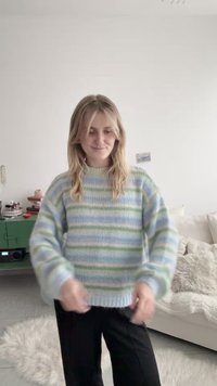 Νεαρή γυναίκα που φοράει μπλε και πράσινη ριγέ πουλόβερ και μαύρο παντελόνι, στέκεται σε έναν ζεστό και άνετο χώρο καθιστικού με έναν καναπέ και μια πράσινη κονσόλα.