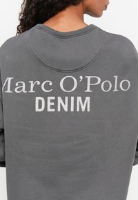Grijze sweatshirt met geribbelde halslijn, waarop "Marc O'Polo" en "DENIM" in grote witte letters op de achterkant staat. Zachte, textuurrijke stof.