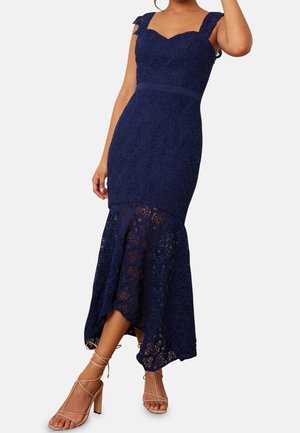 Vrouw draagt een marineblauwe kanten zeemeerminjurk met geschulpte bandjes en nude gestripte blokhakken, poserend met één arm omhoog.