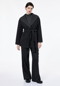 Abrigo de mezcla de lana oscura con cuello ancho, cinturón para ajustar y grandes bolsillos delanteros, combinado con pantalones anchos oscuros y un suéter texturizado.