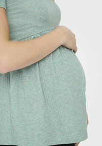 Maternitātes tops gaiši zaļā ribota auduma, ar savilktu jostas vietu, pielāgotu krūšu daļu un īsām piedurknēm. Uzsvars uz vēdera apgabalu, kur uz tā atpūšas roka.