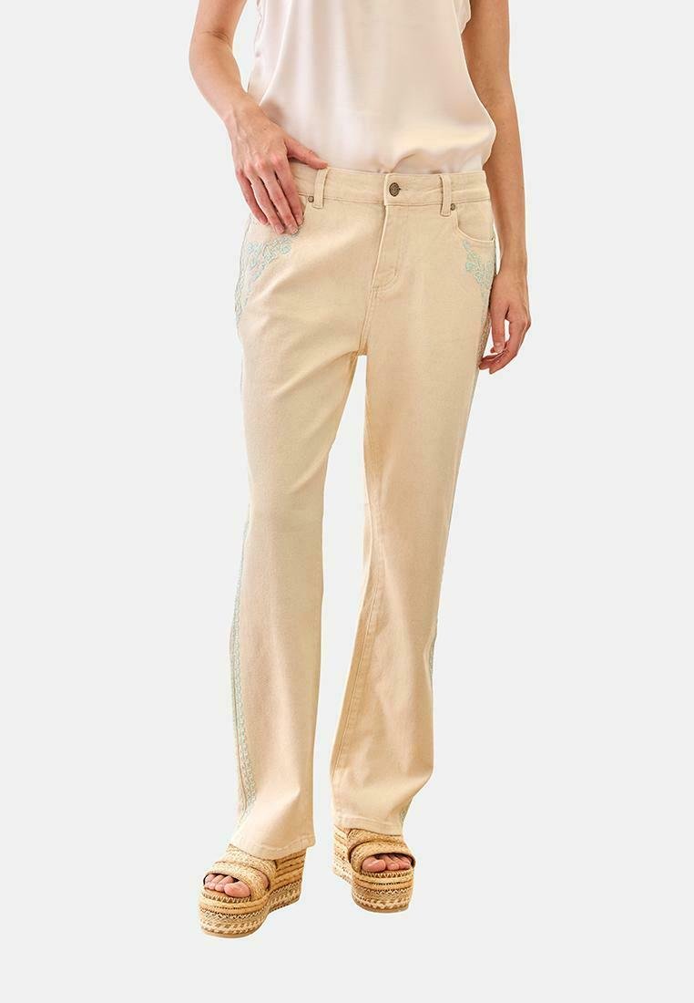 Mujer con pantalones rectos de color crema con bordados sutiles en los bolsillos y los laterales, combinados con sandalias de plataforma beige, de pie sobre fondo blanco.