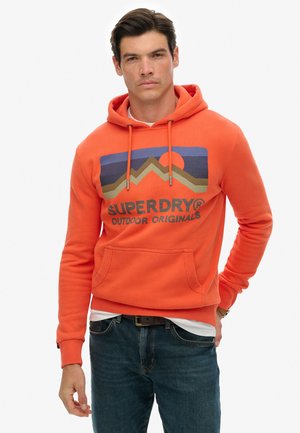 Oranje hoodie met een trekkoord, voorzak en een grafisch ontwerp met bergen en een zon. Gecombineerd met blauwe jeans.
