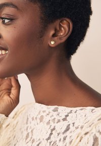 Goldene Ohrstecker mit strukturierter Oberfläche, getragen von einer Person mit kurzem, natürlichem Haar. Der Hintergrund ist neutral und sanft verschwommen.