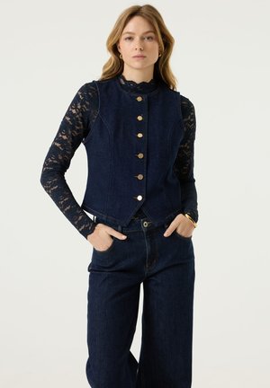 Vrouw met lang lichtbruin haar draagt een navykleurig spijkervest, kanten top met lange mouwen en wijde spijkerbroek, handen in de zakken, neutrale achtergrond.