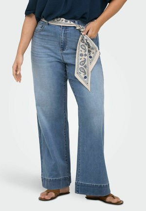 Donna che indossa jeans azzurri a gamba larga, cintura foulard bianca e blu navy con fantasia, top blu navy e sandali piatti marroni.