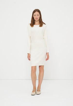 Femme aux cheveux bruns et raides portant une robe blanche à manches longues, longueur genoux, et des chaussures plates beiges, debout sur un fond blanc.