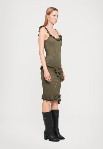 Rochie ajustată de culoare verde măsliniu, cu accente volan la decolteu și la tiv, având un finisaj texturat. Bocanci negri cu toc, până la genunchi, completează ținuta.