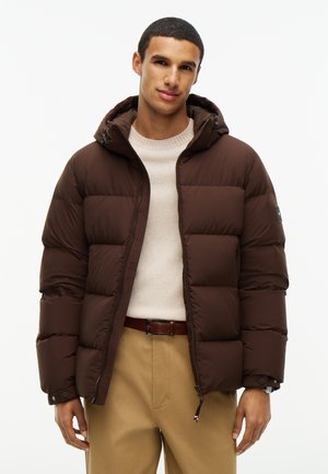 Joven con una chaqueta acolchada marrón con capucha, suéter crema y pantalones beige, de pie frente a un fondo blanco liso.