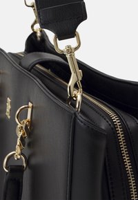 Sac à main en cuir noir avec une fermeture éclair dorée, une chaîne et un détail de fermoir dorés, et une bandoulière noire amovible avec un crochet doré.