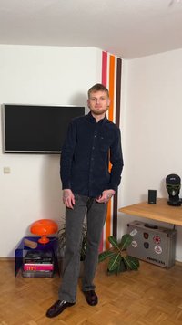 Tmavě modrá manšestrová košile, tmavé džíny, leštěné boty. Má tetování, stojí v místnosti s moderním zařízením, rostlinami a televizí.