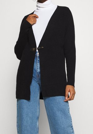 Osoba nosící černý kardigan s otevřeným předním dílem a sponou, bílý rolák a modré džíny s vysokým pasem na jednobarevném pozadí.