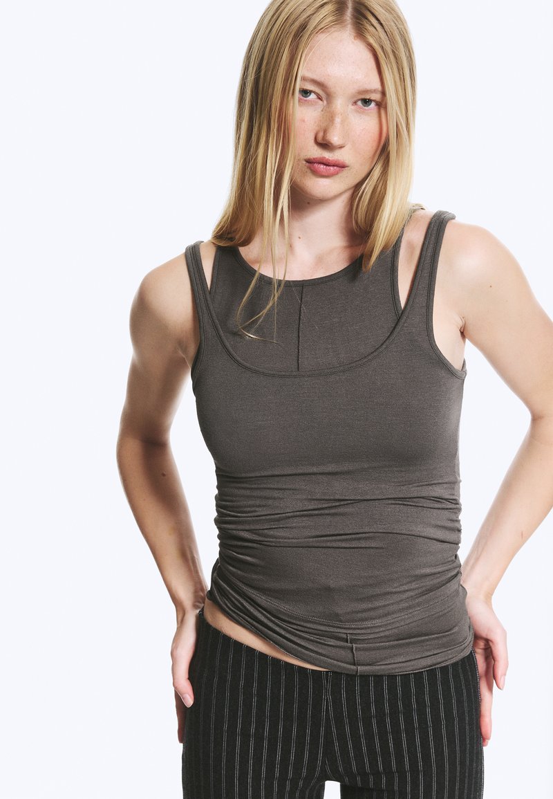 Femme aux cheveux blonds lisses portant un débardeur gris foncé ajusté et un pantalon noir à rayures fines, debout les mains sur les hanches devant un fond uni.