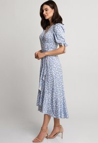 Niebieska kwiatowa sukienka w stylu kopertowym z bufkowymi rękawami, do kolan, z miękkiego materiału, z wiązanym w talii pasem i asymetrycznym dołem; zestawiona z nagimi sandałami na obcasie.