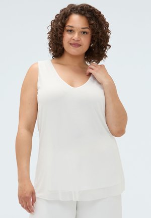 Donna dai capelli ricci che indossa un top bianco senza maniche con scollo a V, che tocca delicatamente il colletto, su uno sfondo chiaro e uniforme.