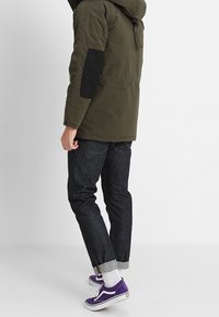 Olivově zelená mikina s kapucí a černými loketními záplatami, spárovaná s tmavými džínovými kalhotami a fialovými teniskami, ukazujícími ohrnuté manžety a uvolněný střih.