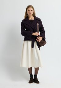 Kort lila peacoat med dubbelknäppt front och bälte, matchat med en vit midikjol. En strukturerad brun väska och mörka skor fulländar looken.
