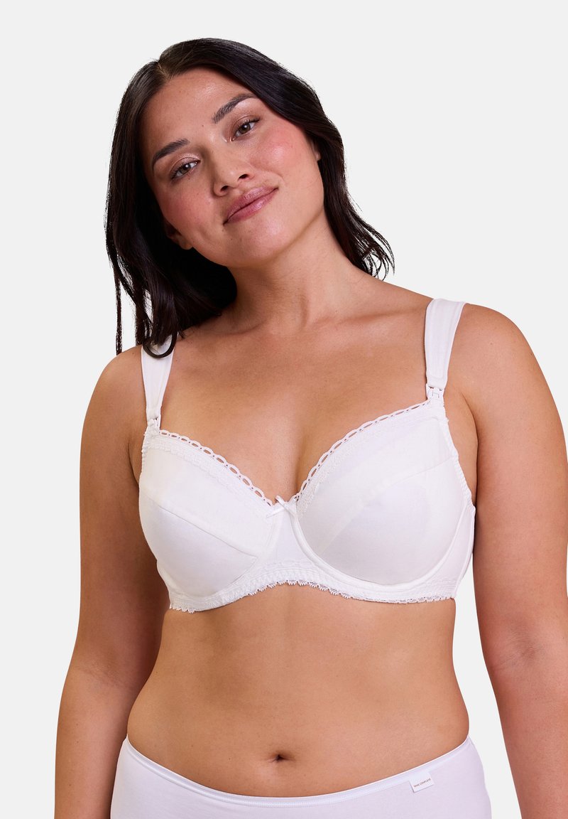 Woman with medium skin tone and dark hair wearing a white padded bra and matching bottoms, smiling slightly against a plain background.
