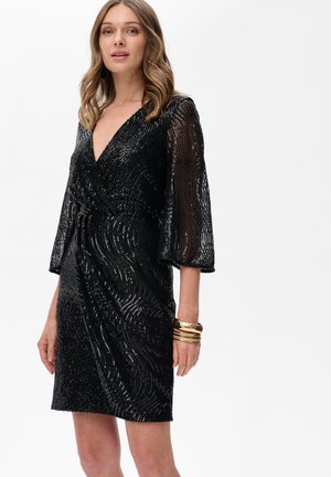 Kobieta w czarnej sukience z cekinami do kolan, z przezroczystymi długimi rękawami i głębokim dekoltem w kształcie litery V, ozdobiona wieloma złotymi bransoletkami.