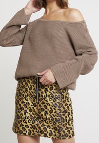 Hnědý pletený svetr s off-shoulder designem a žebrovanou strukturou, kombinovaný se žlutou sukní z umělé kůže s leopardím vzorem a detaily na zip.