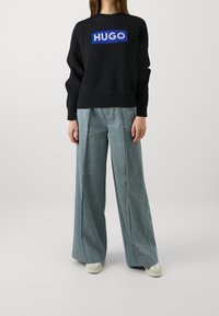 Schwarzer Strickpullover mit blauem "HUGO"-Logo, kombiniert mit hellblauen weit geschnittenen Hosen. Das Outfit zeichnet sich durch eine lockere Passform und ein geripptes Muster aus.