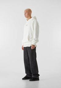 Felpa bianca con tasca a marsupio, logo nero, indossata sopra una maglietta bianca a maniche lunghe. Abbinata a jeans neri a vestibilità rilassata e sneakers nere.