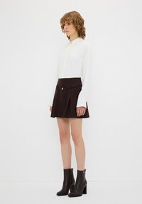 Chemisier blanc à boutons associé à une jupe plissée brun avec une boucle dorée et une taille cintrée ; le mannequin porte des bottines brunes.