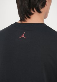 Must musta t-särk, millel on taga roosa Jordan logo. Lühikesed varrukad ja lihtne, klassikaline ümar kaelus. Sile tekstuur.