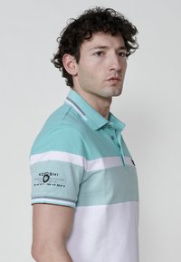 Törökzöld és fehér csíkos polo póló gallérral, logóval és szöveggel az ujjon, strukturált anyagból készült.