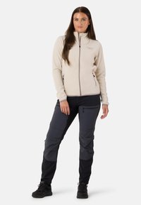 Vaaleanbeige fleece-takki korkealla kauluksella ja vetoketjulla; yhdistetty tummanharmaisiin vaellushousuihin, joissa on taskuja ja mustia yksityiskohtia.