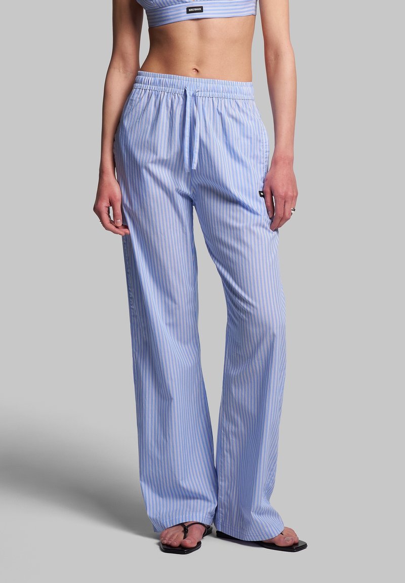 Femme portant un pantalon large à fines rayures bleu clair et blanc avec un cordon à la taille et des sandales noires style tongs sur fond gris.