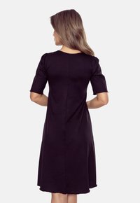 Kobieta z długimi włosami w czarnej sukience, widziana od tyłu na jasnoszarym tle. Sukienka ma krótkie rękawy i luźny krój.