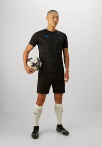 Juodas sportinis džersis su zigzago raštu, derinamas su juodomis šortais, baltais kojiniais ir juodais sportbačiais. Laiko juodai baltą futbolo kamuolį.
