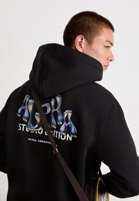 Felpa nera con cappuccio e ampia grafica lucida "ALPHA" in blu e bianco. Presenta un cappuccio con cordino e una tracolla marrone.