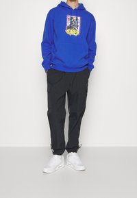 Blå hoodie med grafiskt tryck som föreställer en hund och texten "OBEY"; svarta cargobyxor; vita sneakers med synlig luftkudde-detalj.